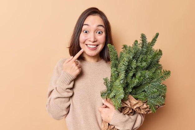 La donna asiatica positiva indica il sorriso a trentadue denti esprime emozioni positive abbraccia il bouquet di rami di abete ritorna dal mercatino di Natale si prepara per le vacanze invernali isolate sopra il muro marrone