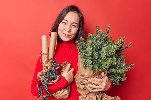 La donna asiatica soddisfatta sorride con gioia tiene gli articoli per le vacanze per la decorazione anticipa per il nuovo anno e il Natale indossa pose a collo alto su sfondo rosso vivido. Celebrazione e concetto di preparazione