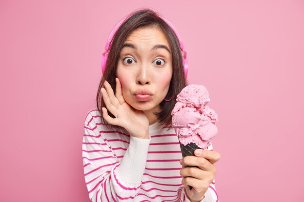 La donna asiatica sorpresa mantiene le labbra arrotondate mangia un delizioso gelato vuole mangiare un dessert congelato freddo indossa le cuffie sulle orecchie maglione a righe casual isolato sul muro rosa. Estate.