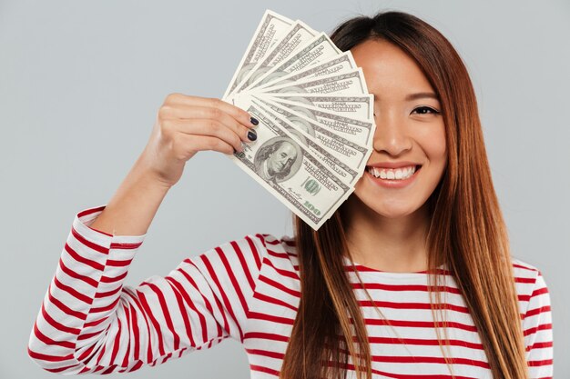 La donna asiatica sorridente in maglione sta coprendo in soldi della metà del fronte e sta esaminando la macchina fotografica sopra backgound grigio