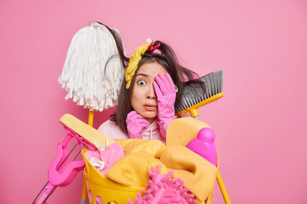 La donna asiatica spaventata tiene la mano sul viso sembra spaventata alla telecamera circondata da strumenti per la pulizia ha paura di iniziare a pulire la stanza molto sporca fa il bucato a casa isolata sul muro dello studio rosa