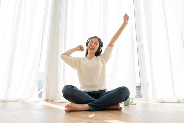 La donna asiatica tranquilla e pacifica si gode il fine settimana con la luce del sole mattutino delle cuffie musicali vicino allo sfondo della finestra di casa