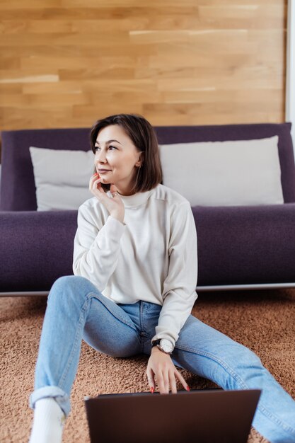 La donna astuta lavora al computer portatile che si siede sul pavimento a casa nel tempo casuale vestita