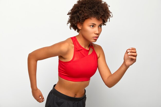 La donna atletica sicura di sé sta in posa da corsa, guarda seriamente al traguardo, dimostra resistenza, indossa un top rosso e pantaloncini, si muove attivamente con le braccia