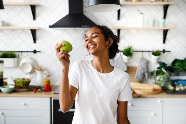 La donna attraente sorridente del mulatto si sta preparando a mordere una mela e sta osservando la mela sulla cucina moderna bianca