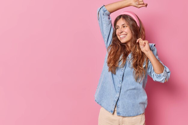 la donna balla con le braccia alzate sorride si muove ampiamente indossa attivamente fascia per capelli e camicia di jeans isolata sullo spazio rosa della copia a sinistra per i tuoi contenuti promozionali