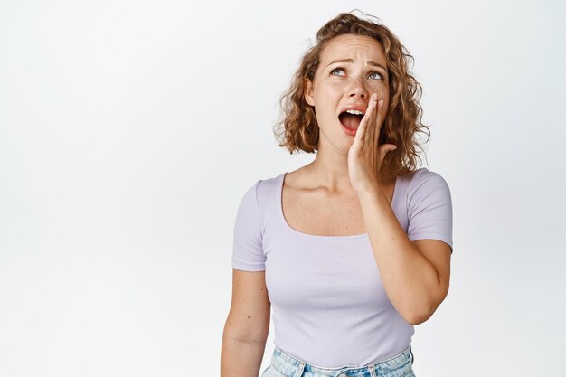 La donna bionda che sbadiglia copre la bocca aperta, guardando annoiata o esausta nell'angolo in alto a sinistra, si erge su sfondo bianco