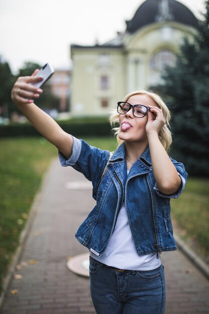 La donna bionda della ragazza di modo alla moda in suite di jeans e occhiali fa selfie sul suo telefono in città al mattino