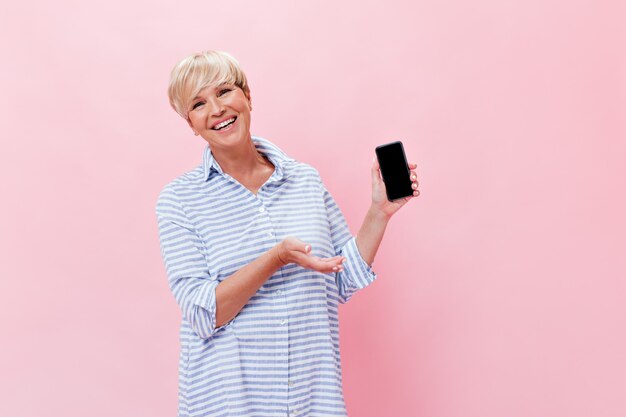 La donna bionda in vestito blu mostra lo smartphone su sfondo rosa