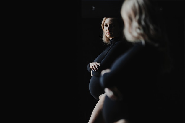 La donna bionda incinta in una tuta nera sta vicino ad uno specchio e esamina il suo riflesso