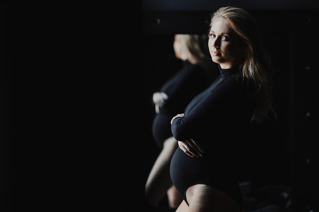 La donna bionda incinta in una tuta nera sta vicino ad uno specchio