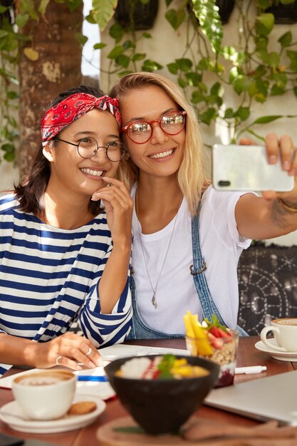 La donna bionda positiva tiene lo smart phone nelle mani, fa la foto del selfie insieme all'amico asiatico, goditi le vacanze estive insieme