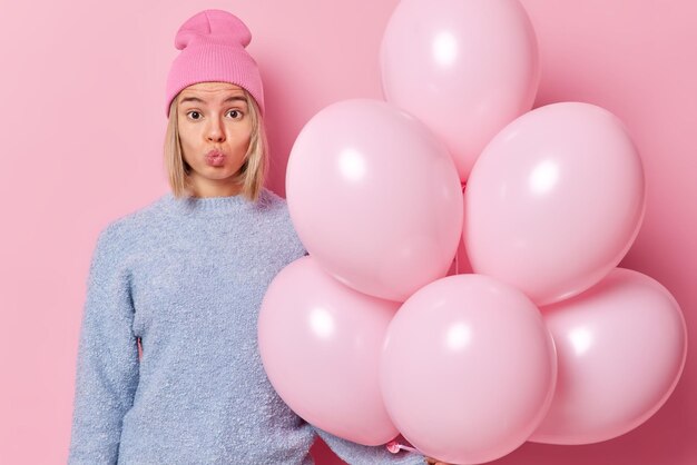 La donna bionda romantica mantiene le labbra arrotondate indossa il cappello e il maglione pronti per la celebrazione tiene un mazzo di palloncini di elio prepara le decorazioni per la festa isolate su sfondo rosa Concetto di vacanze