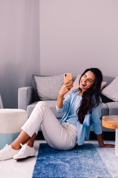 La donna blogger di moda bruna europea in forma si siede sul pavimento nel soggiorno vicino al divano con il telefono