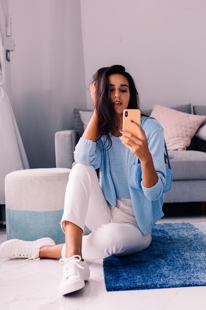 La donna blogger di moda bruna europea in forma si siede sul pavimento nel soggiorno vicino al divano con il telefono