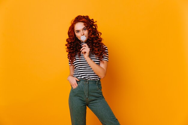 La donna brillante con capelli rossi ondulati lunghi mangia il lecca-lecca e posa in jeans sullo spazio isolato.