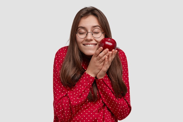La donna bruna soddisfatta prova piacere, tiene una mela gustosa, è di buon umore, indossa abiti rossi