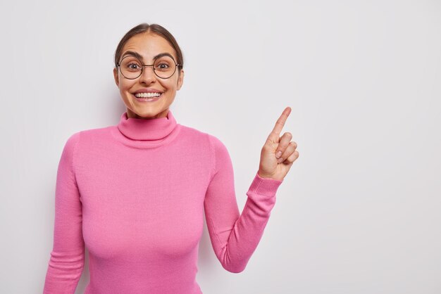 La donna bruna soddisfatta punta il dito indice in uno spazio vuoto consiglia che il prodotto ha un'espressione allegra indossa occhiali rotondi dolcevita rosa casual isolato su sfondo bianco Guarda questo