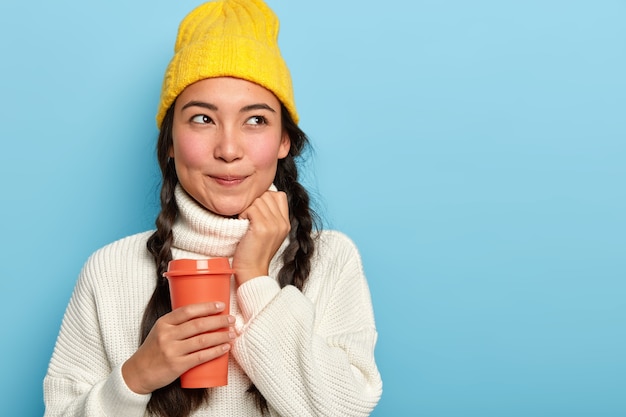 La donna bruna sognante ha un aspetto naturale, indossa un cappello giallo e un maglione bianco, tiene un caffè da asporto, è immersa nei pensieri