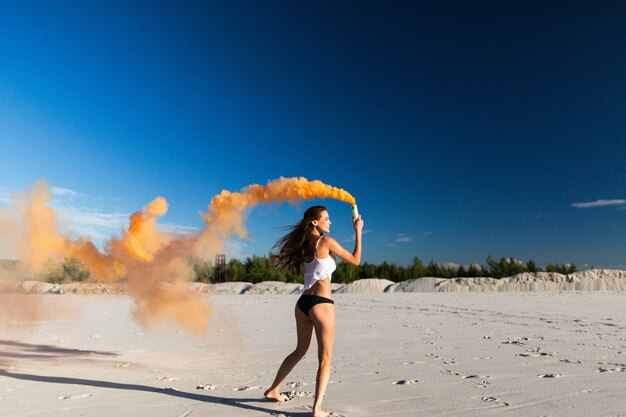 La donna cammina con il fumo arancione sulla spiaggia bianca sotto il cielo blu