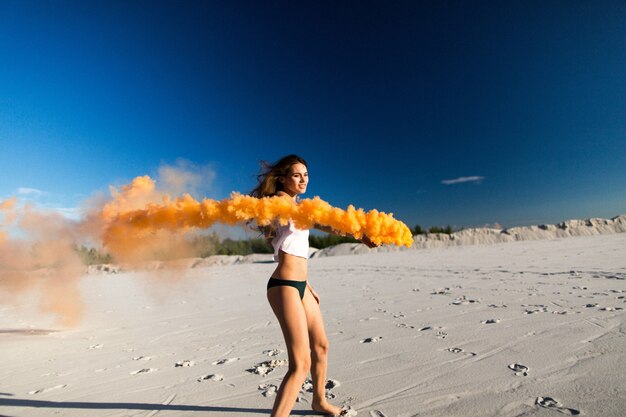 La donna cammina con il fumo arancione sulla spiaggia bianca sotto il cielo blu