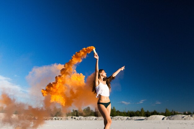 La donna cammina con il fumo arancione sulla spiaggia bianca sotto il cielo blu