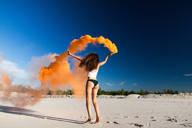 La donna cammina con il fumo arancione sulla spiaggia bianca sotto il cielo blu