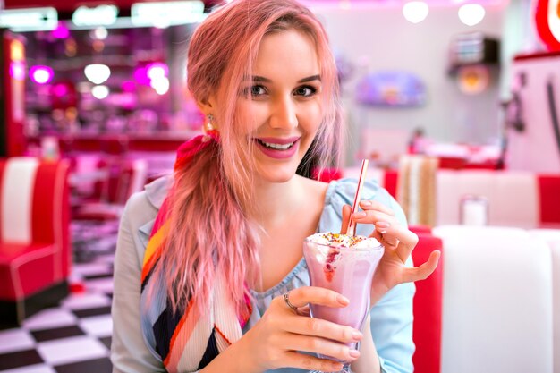 La donna carina e sensuale con i capelli rosa alla moda si gode il suo dolce frullato alla fragola, sorridente, vestito vintage pastello alla moda