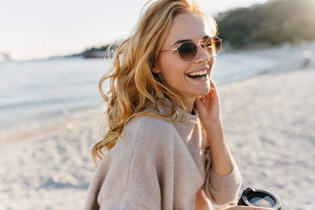 La donna carina ride sinceramente, rilassandosi in spiaggia. Blinde donna in occhiali e maglione tiene la tazza di caffè.