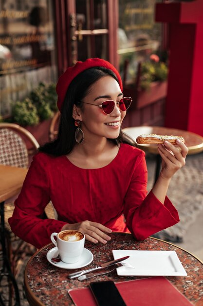La donna castana abbronzata sveglia in vestito rosso alla moda, berretto e occhiali da sole si siede in caffè