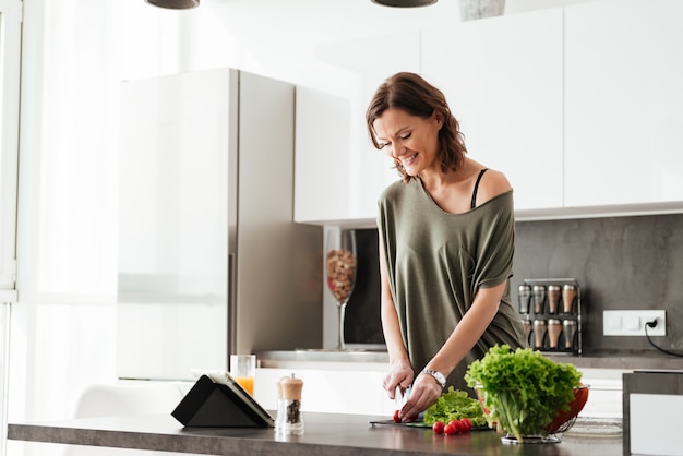 La donna casuale sorridente taglia la verdura dalla tavola sulla cucina e per mezzo del computer della compressa