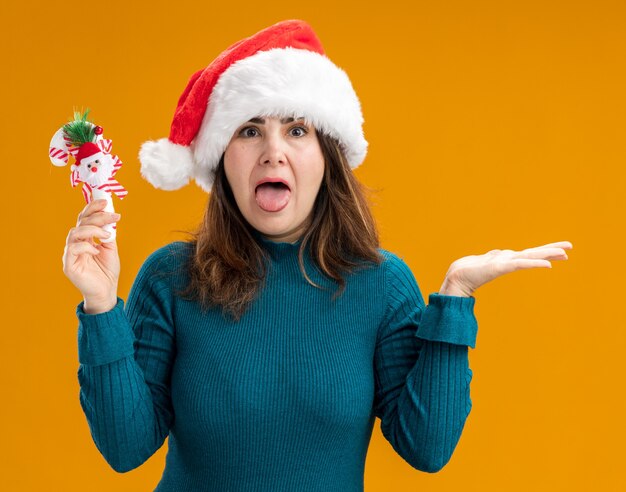 La donna caucasica adulta infastidita con il cappello della Santa sporge la lingua e tiene il bastoncino di zucchero isolato su fondo arancio con lo spazio della copia