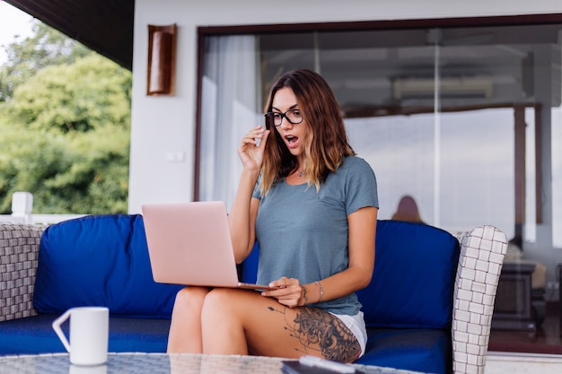 La donna caucasica felice fa il lavoro a distanza sul computer portatile nel posto comodo di casa che beve il tè