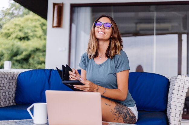 La donna caucasica felice fa il lavoro a distanza sul computer portatile nel posto comodo di casa che beve il tè
