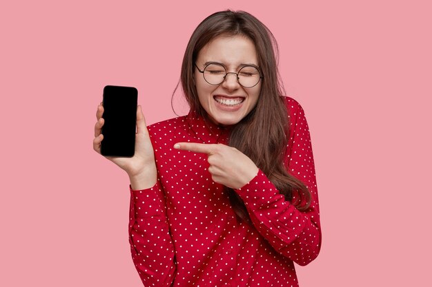 La donna caucasica ottimista indica il nuovo smartphone per la pubblicità, mostra lo schermo vuoto, ama il gadget multifunzionale
