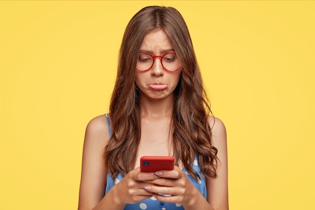 La donna caucasica scontenta ha un'espressione offensiva, increspa le labbra inferiori per il dispiacere, usa il moderno telefono cellulare