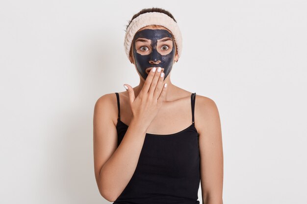 La donna caucasica sorpresa emozionale guarda con espressione scioccata, sta contro il muro bianco, pulisce la pelle con la maschera cosmetica al fango.