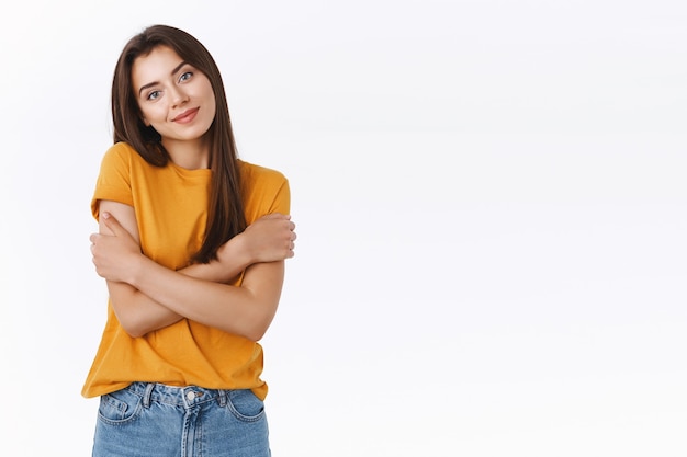 La donna caucasica tenera e femminile pensa che sia il momento di indossare vestiti caldi, l'inverno in arrivo, abbracciarsi con le mani incrociate e un sorriso sensuale, inclinare la testa, guardare la fotocamera gioiosa e civettuola, sfondo bianco