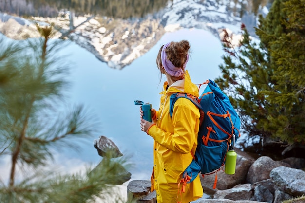 La donna che fa un'escursione si ferma vicino al lago in montagna, trasporta la schiena, tiene un thermos di bevanda calda, esplora qualcosa di nuovo