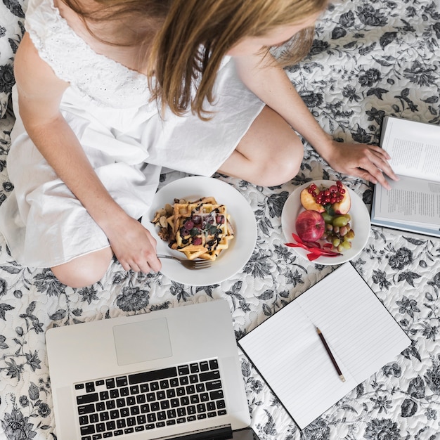 La donna che si siede sul letto floreale con la cialda e fruttifica libro di lettura della prima colazione