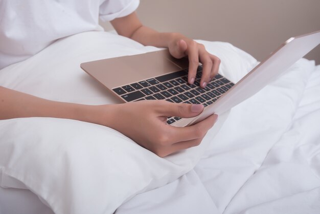 La donna che usando la seduta sorridente del computer portatile sul letto a casa ha svegliato di mattina