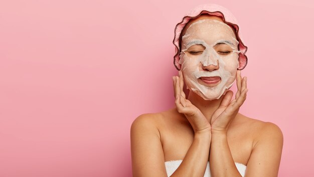 La donna cinese soddisfatta gode della procedura cosmetica, ha una maschera facciale di carta naturale sulle guance, avvolta in un asciugamano, indossa la cuffia da bagno, ha gli occhi chiusi, isolato su un muro rosa con spazio libero per il tuo annuncio