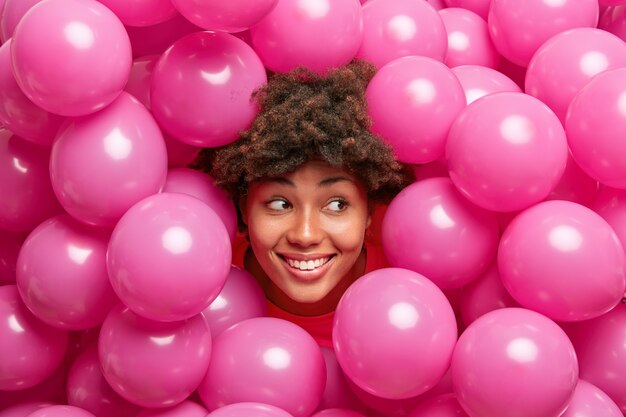 la donna con i capelli ricci e folti sorrisi sembra ampiamente a destra ha un umore festivo pone intorno a palloncini rosa elio gonfiati