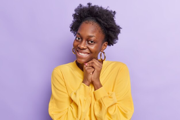 la donna con i capelli ricci naturali tiene le mani unite vicino al viso sorride con i denti si sente toccata indossa un maglione giallo casual isolato su viola