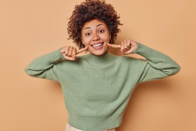 la donna con i capelli ricci punta al sorriso mostra denti bianchi perfetti vestiti con un maglione casual isolato sul beige essere di buon umore. Espressioni felici del volto umano