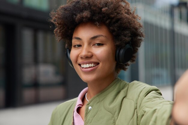 la donna con i capelli ricci sorride ascolta positivamente la traccia audio o la composizione preferita tramite le cuffie wireless ha pose di umore ottimista all'aperto.