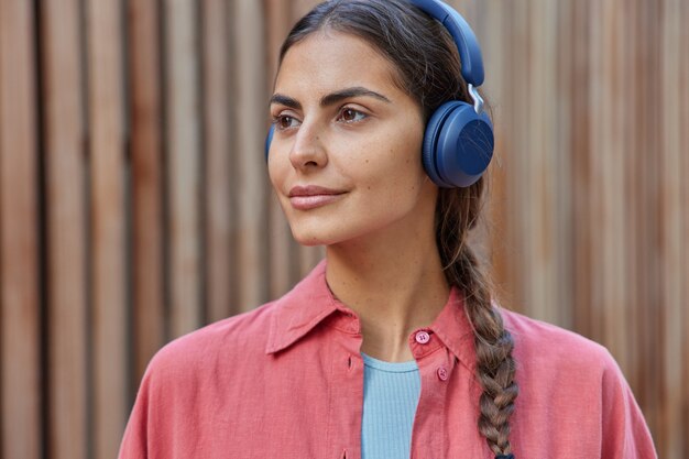 la donna con il codino ascolta la musica tramite le cuffie wireless gode di una playlist di musica preferita positiva focalizzata lontano vestita in camicia pone su sfocato