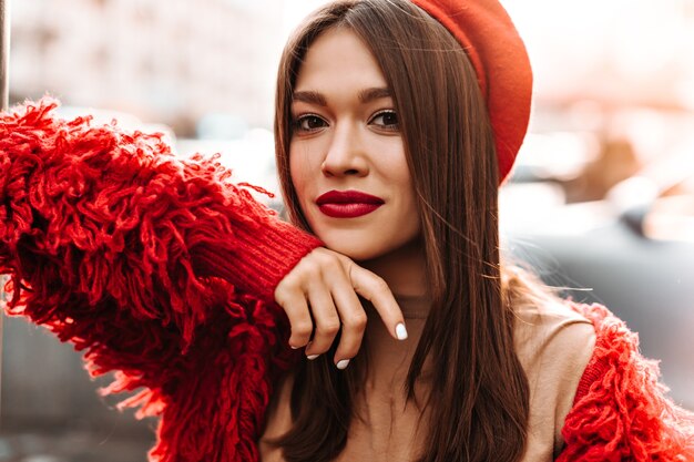 La donna con il manicure bianco e le labbra rosse sta esaminando la macchina fotografica sullo sfondo della strada. donna vestita di berretto rosso e giacca di lana appoggiata alla finestra.