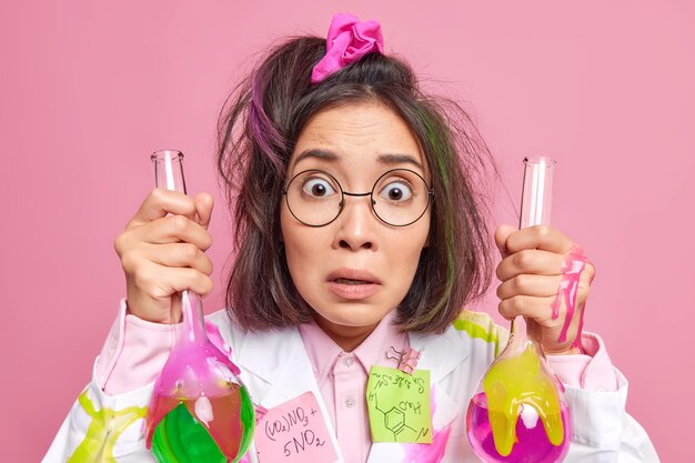 la donna conduce ricerche scientifiche vestita con camice medico tiene bicchieri di vetro con liquido colorato sorpresa dai risultati dell'esperimento indossa occhiali rosa