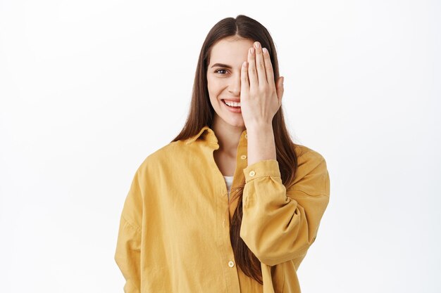 La donna copre una metà del viso e sorride alla macchina fotografica, prima dopo il concetto, con il trucco naturale nudo, in piedi in camicia oversize elegante gialla, sfondo bianco